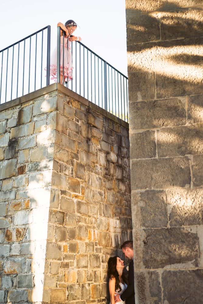 a young girl dressed as a flower girl is looking down from the stair to the newlyweds that are under the stairs kissing each other in a romantic way